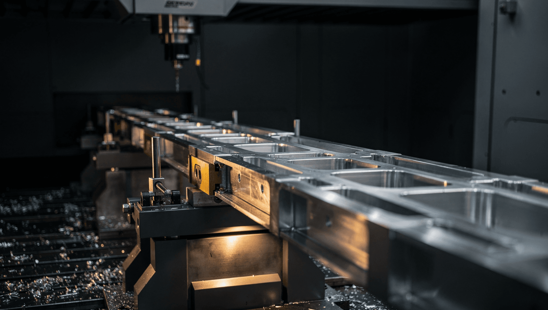 A long aluminum aerostructure spar clamped on its machining fixture beneath a gantry mill head, rows of machined pockets receding down its length under cool light, one warm brass highlight at the fixture clamp, and fresh metal swarf scattered across the near-black bay.