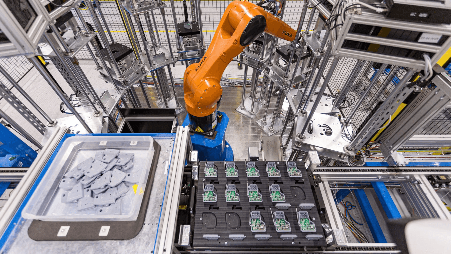 A saturated-orange six-axis industrial robot arm centered in a wire-mesh caged assembly cell framed in aluminum extrusion, a clear tote of grey moulded parts staged at lower left and a black nest matrix holding rows of green circuit boards at lower right.