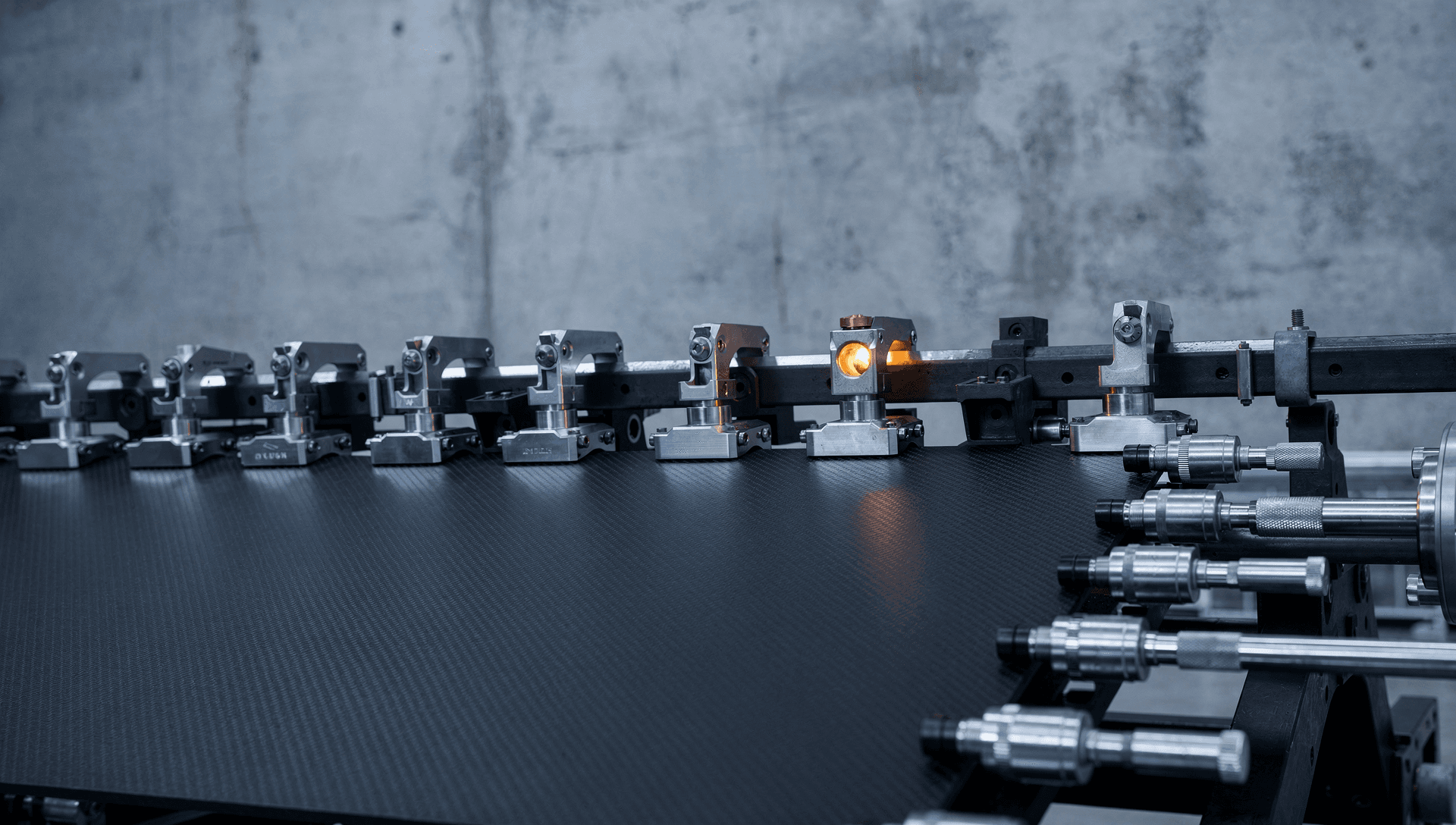 A composite panel section fixtured and drilled in a large integration jig in a US aerospace assembly area, drill templates and clamps along the edge, cool blue-grey industrial light with a warm brass highlight on a drill-fixture bushing.