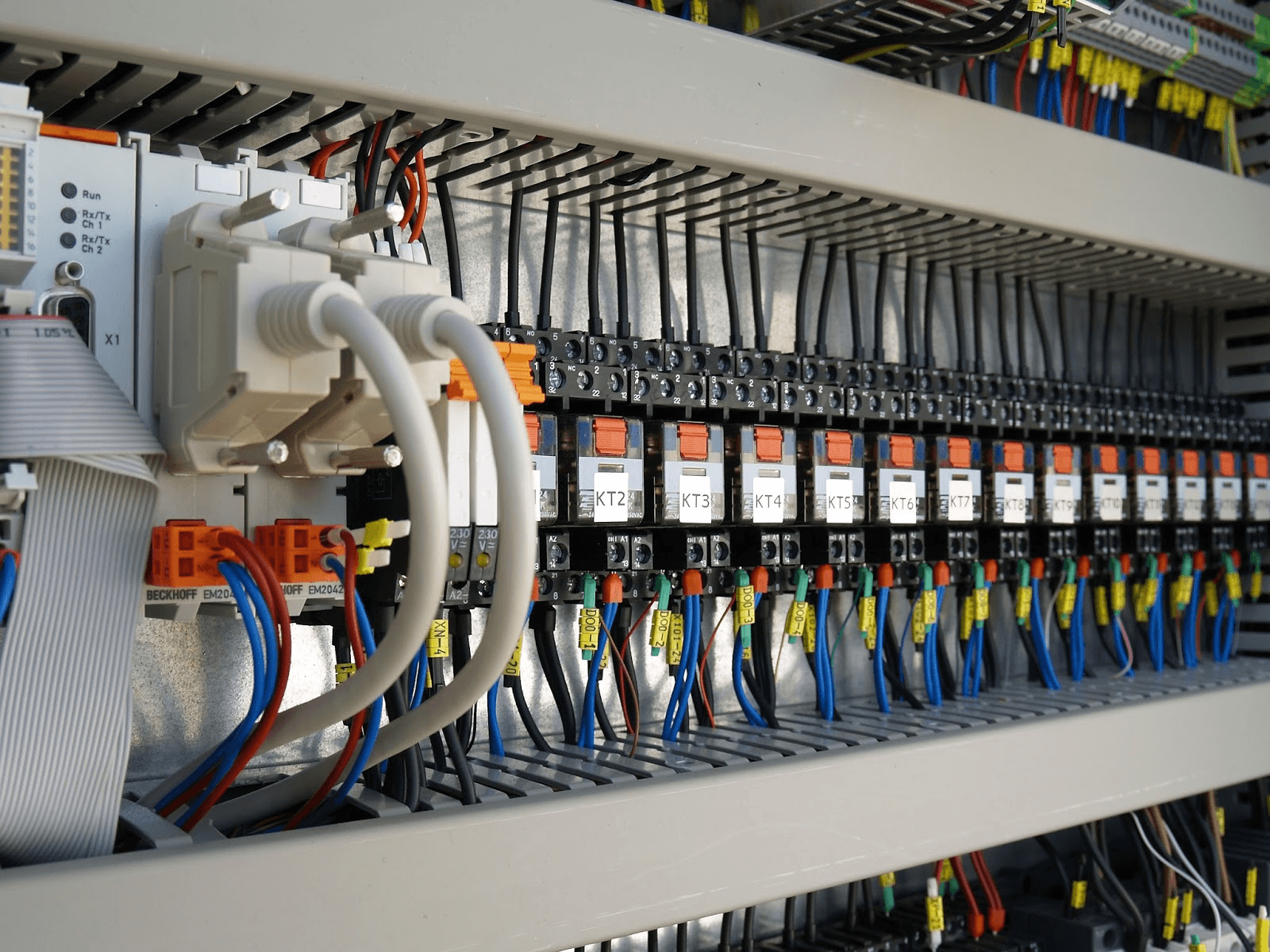 A receding rank of identical labeled relays over numbered terminal strips inside an open control cabinet, every conductor ferruled, tagged, and dressed into the wire duct, with a compact PLC bus coupler anchoring the row.
