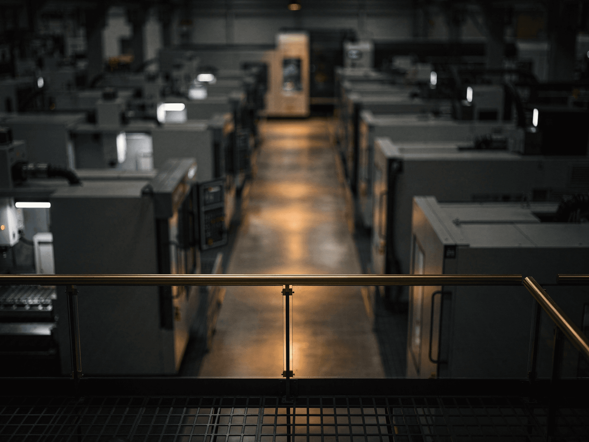 An elevated view from a steel mezzanine walkway over a darkened production floor, one aisle below lit in a long pool of warm light with CNC machines receding in rows into shadow, the handrail crossing the lower frame with a single warm brass glint on its top rail.