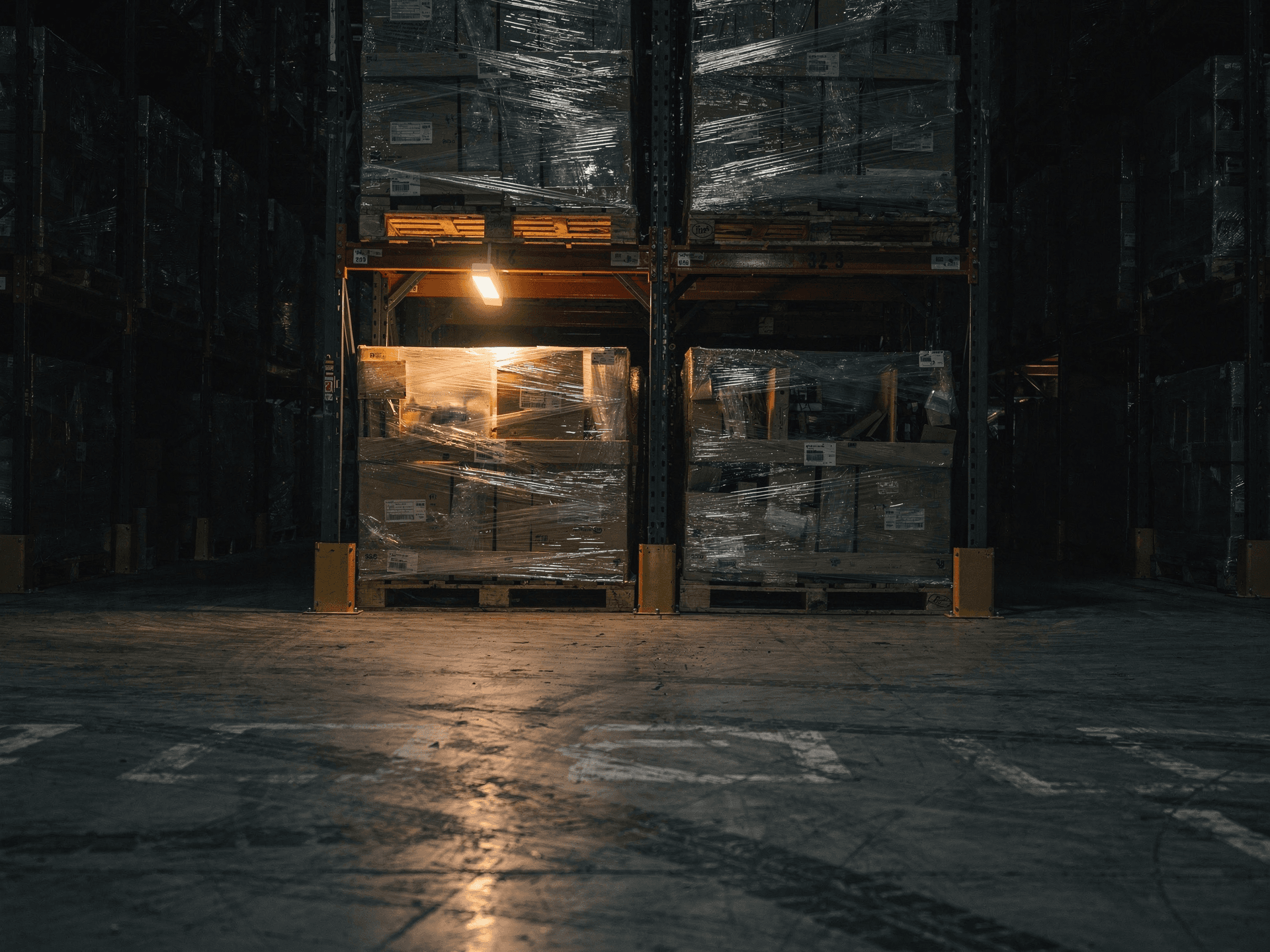 Stretch-wrapped pallets of work-in-process stacked two levels high in deep pallet racking in a dark warehouse corner, the plastic wrap catching a single overhead bay light so the trapped inventory glows against the near-black aisle.